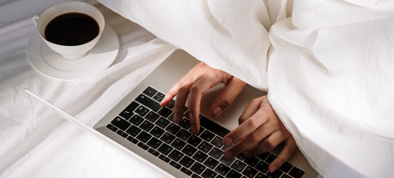 Someone typing under sheets with a cup of coffee.
