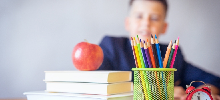 A blurred photo of a child in the background and some coloring pencils and an apple on top of books