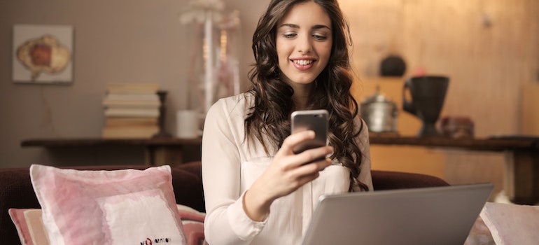 A woman looking at a phone and smiling with a laptop open in front of her