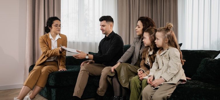 a realtor in a brown suit handing papers to a family of four sitting on a black velvet couch