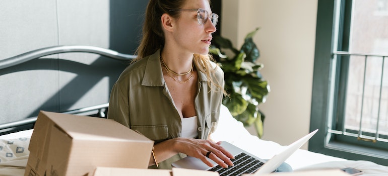 Woman with glasses sitting on her bed with her laptop and cardboard boxes searching for best cross country movers Toms Rive, New Jersey