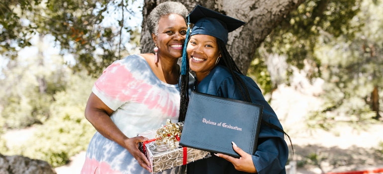 A woman who graduated posing with a diploma in her hand next to her mom holding a wrapped gift