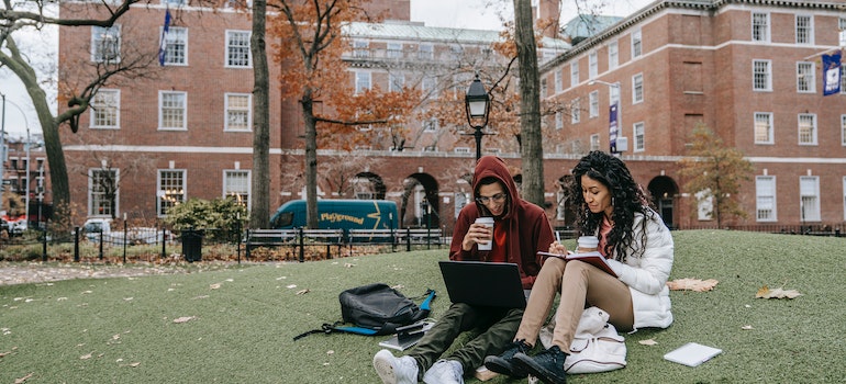 Two students sitting on a lawn on campus in front of a university building looking at their papers