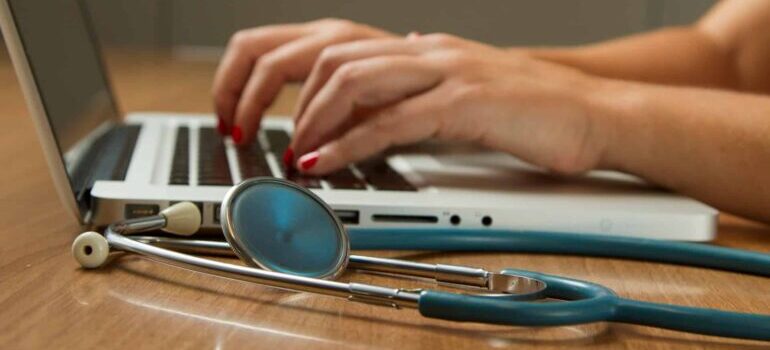 Hands typing on laptop keyboard, stetoscope next to it